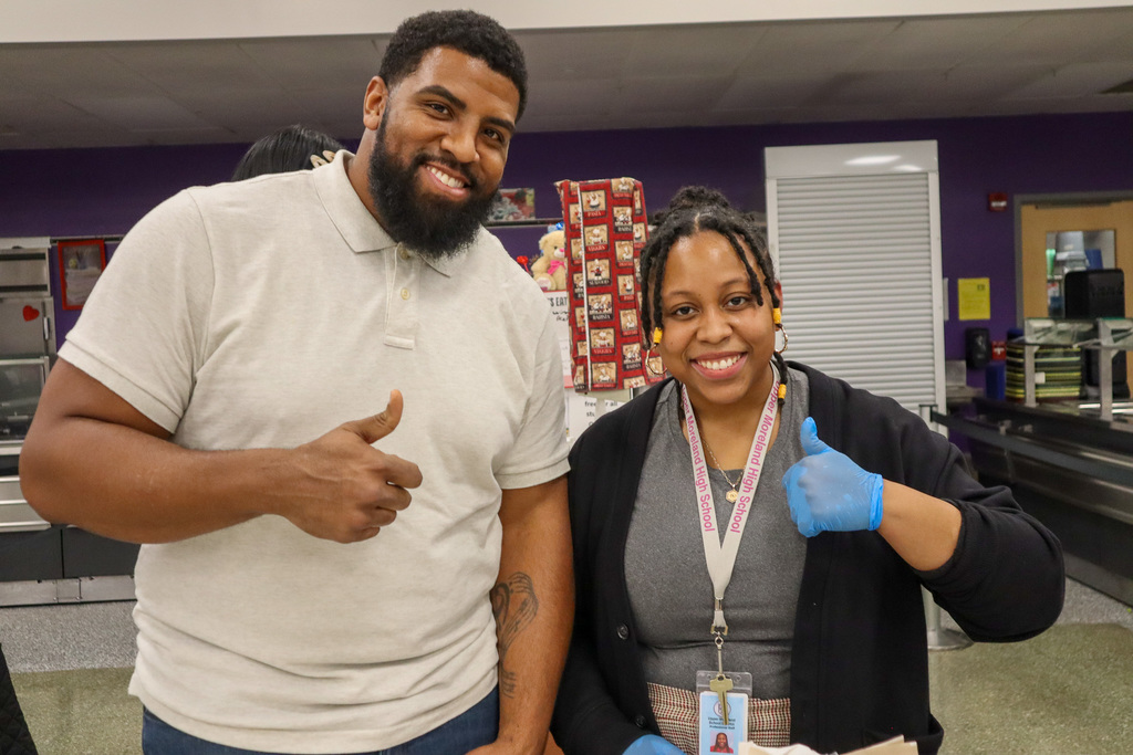 Two people giving thumbs-up gestures indoors, one wearing a light gray polo, the other with a cardigan and blue gloves.