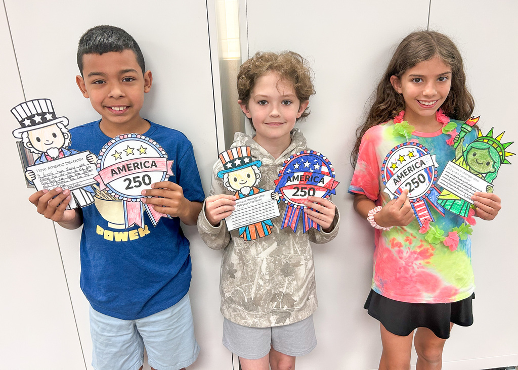 Three children holding craft projects related to "America 250." The first child on the left is a boy in a blue shirt, the middle child is a boy in a light hoodie, and the girl on the right is wearing a tie-dye shirt and a flower lei.
