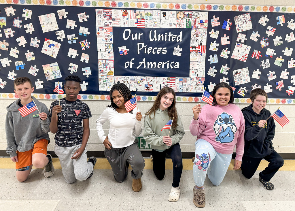 A group of six children kneel in front of a bulletin board with the message "Our United Pieces of America," holding small American flags and smiling.