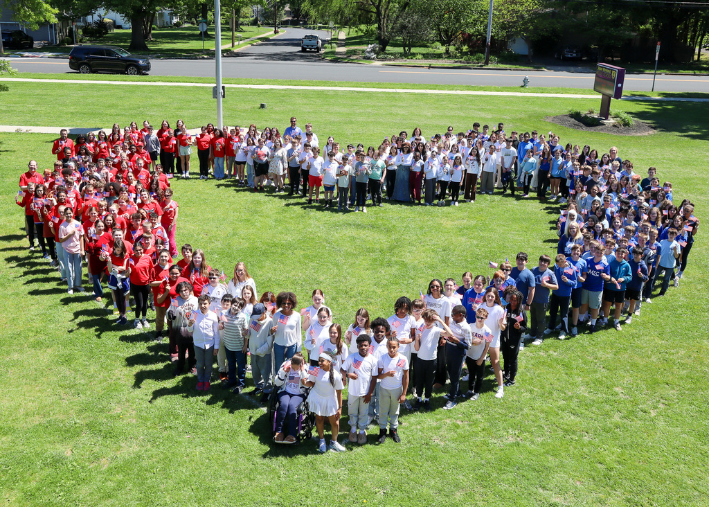 A large group of people forming a heart shape on a grassy field, wearing red, white, and blue t-shirts, with some smiling and waving.