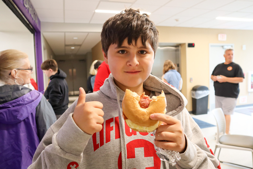 A young boy with dark hair holds a hot dog and gives a thumbs-up in a busy indoor setting.