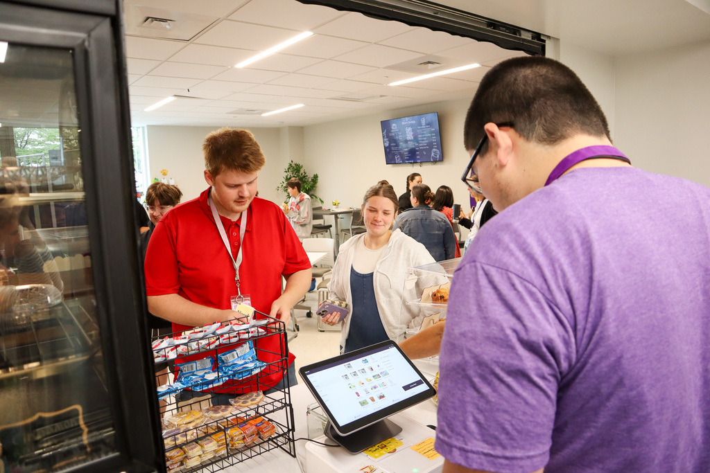 Customers interact at a food service counter as staff serve snacks in a modern, busy environment, with a digital menu board in the background.