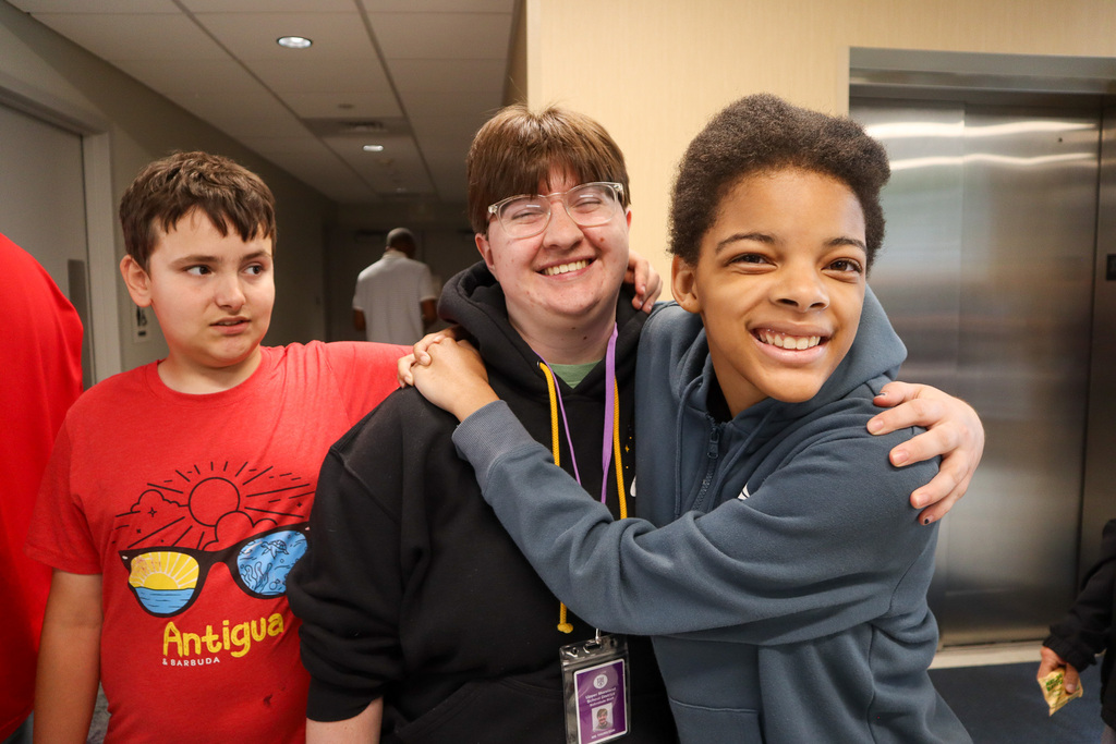 Three young individuals are smiling in a hallway; one is wearing a red t-shirt, the center person has glasses and is hugging the third person, who is embracing them joyfully.