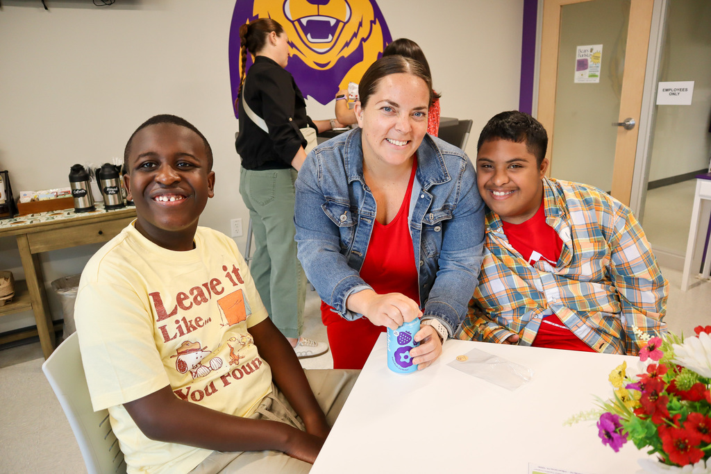 Three individuals at a table: a smiling boy in a yellow shirt, a woman in a red top and denim jacket opening a container, and another boy in a red shirt with a plaid shirt.