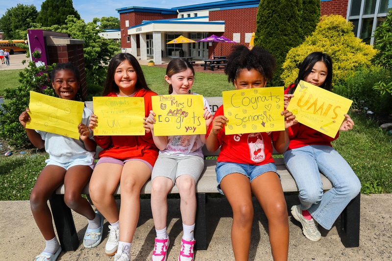 Group of five UMIS students hold yellow signs while sitting on a bench