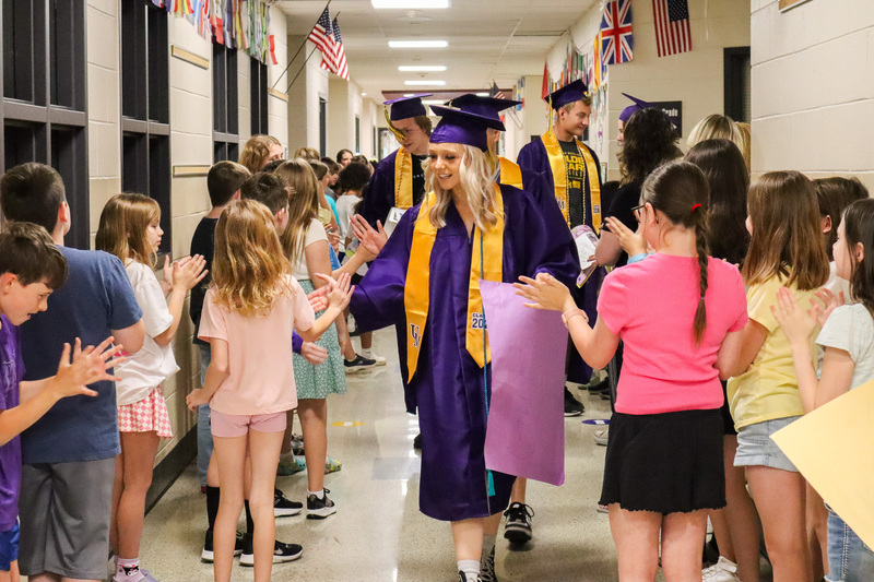 Senior student in graduation attire walks through UMIS hallway and high fives younger students