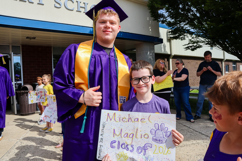 Senior student wearing graduation attire stands next to Primary School student holding a sign with the Senior's name on it