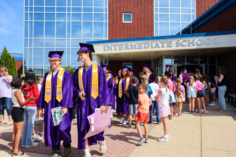 Seniors in graduation attire walk out of the UMIS building through a line of younger students and staff members