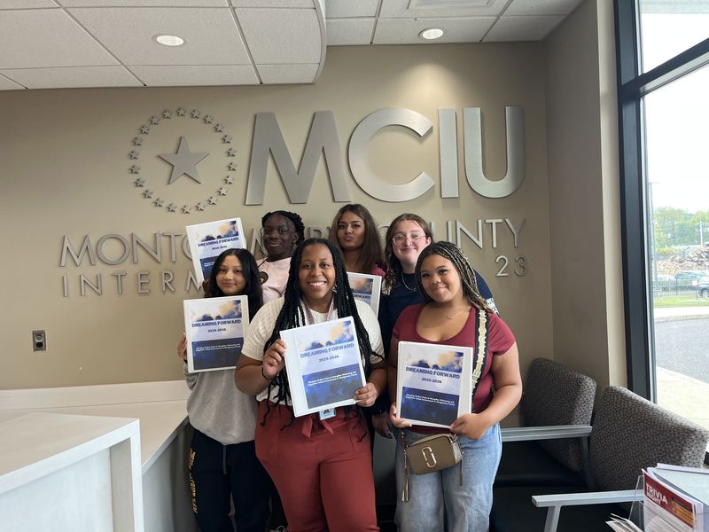 A group of six people holding certificates in front of a sign that reads "MCIU Montgomery County Intermediate Unit 23."