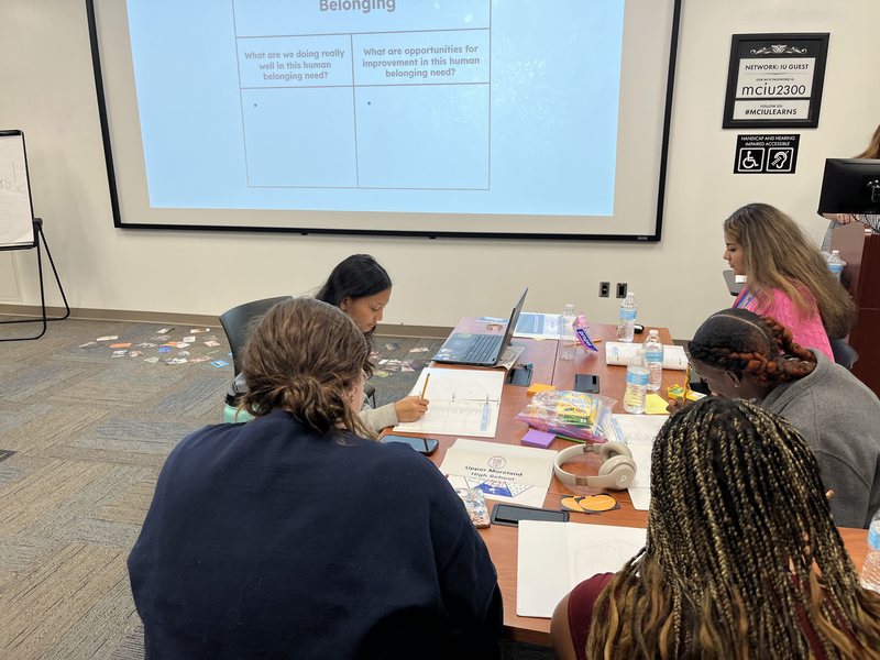 A group of students seated at a table with a presentation projected on the wall.
