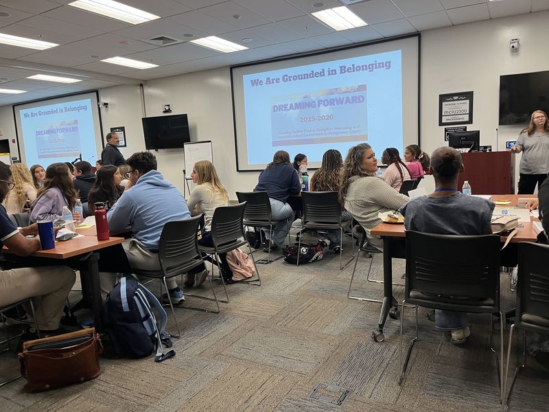 Classroom with people seated at tables, watching a presentation on two large screens.