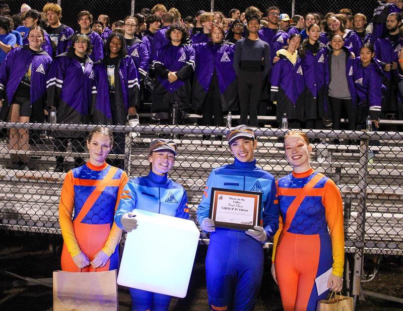 Performers in colorful costumes pose with a certificate and a glowing cube in front of a crowd at a nighttime event.