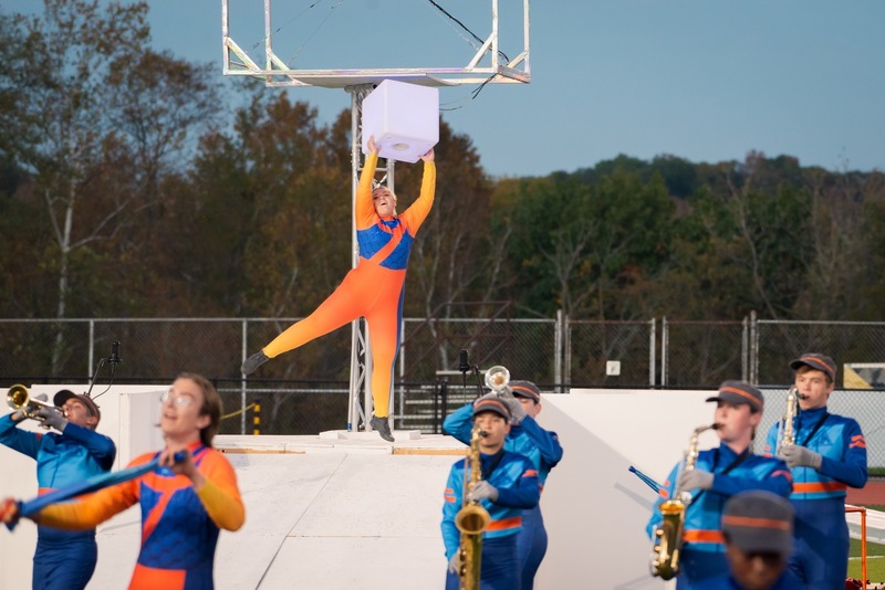 A performer in an orange and blue costume holds a white cube on a platform, surrounded by marching band members.