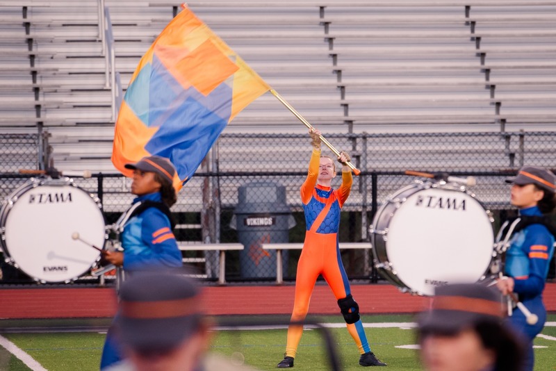 A performer in an orange and blue uniform waves a large flag with geometric patterns on a sports field.
