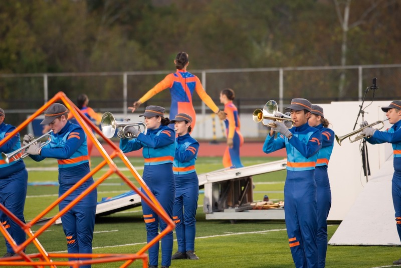 Marching band musicians in blue and orange uniforms play brass instruments on a field.