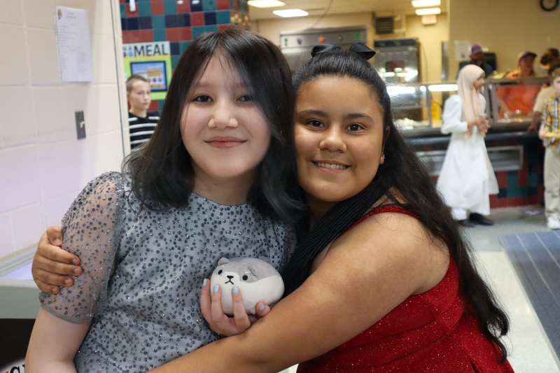 Two people smiling and hugging in a cafeteria setting, one holding a plush toy.