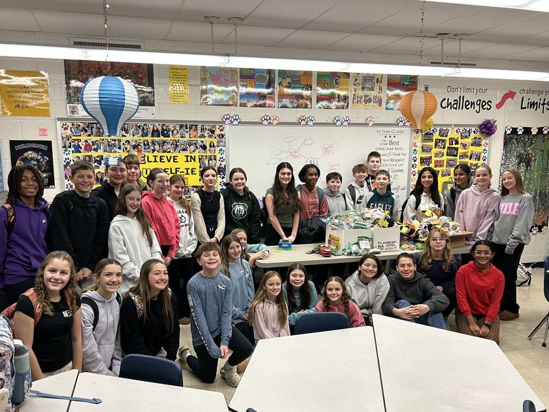 A group of smiling students gathered around a table loaded with packages in a decorated classroom.