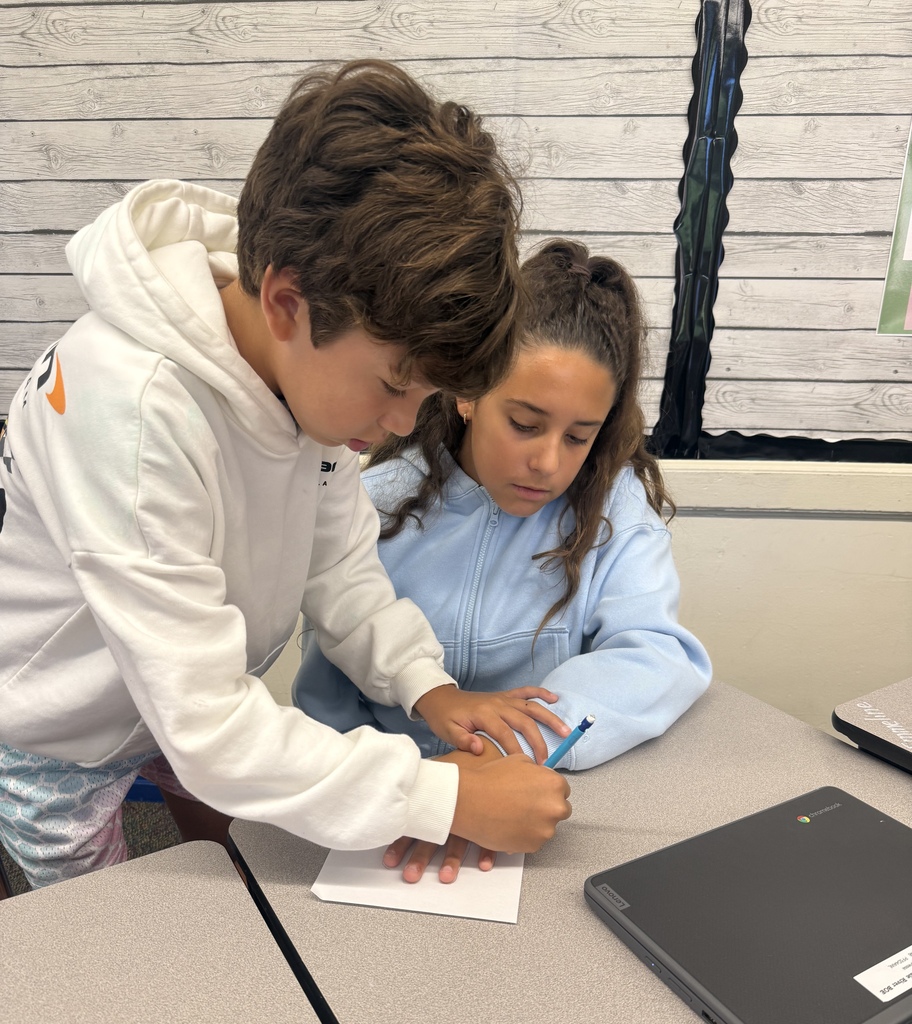 2 students work on an activity on a classroom desk. One student is tracing another students hand. 