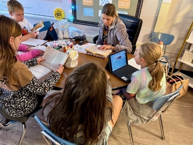 A small group of elementary students sits around a classroom table participating in a reading activity. Several students hold open copies of a novel while another uses a tablet. An adult seated at the table reads from an open book and helps guide the discussion. The classroom contains school supplies, notebooks, and educational posters, creating a collaborative learning environment focused on literacy and reading comprehension.