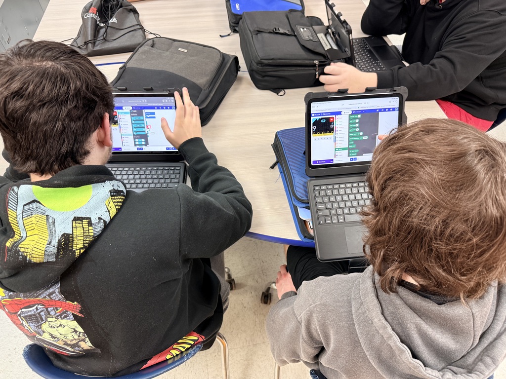 Overhead view of several high school students seated around a classroom table using iPads with keyboard cases. The devices display coding and programming activities as students collaborate on digital projects. Backpacks and laptops are visible on the table, highlighting a technology-rich learning environment focused on developing real-world skills, problem-solving, and digital literacy in a public school classroom.