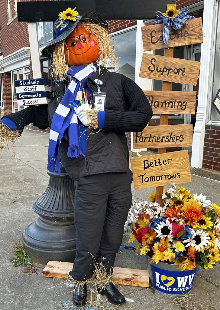 A fall-themed scarecrow display stands on a downtown sidewalk in front of a brick building. The scarecrow has a pumpkin head, straw hair, glasses, a black hat decorated with a sunflower, a blue-and-white scarf, and a school ID badge. It holds a sign reading “Students, Staff, Community, Success.” Beside the scarecrow, wooden signs list “Educate,” “Support,” “Planning,” “Partnerships,” and “Better Tomorrows.” A flower pot filled with colorful autumn flowers displays an “I ❤️ WV Public Schools” logo. The display is decorated with sunflowers, straw, and fall foliage.