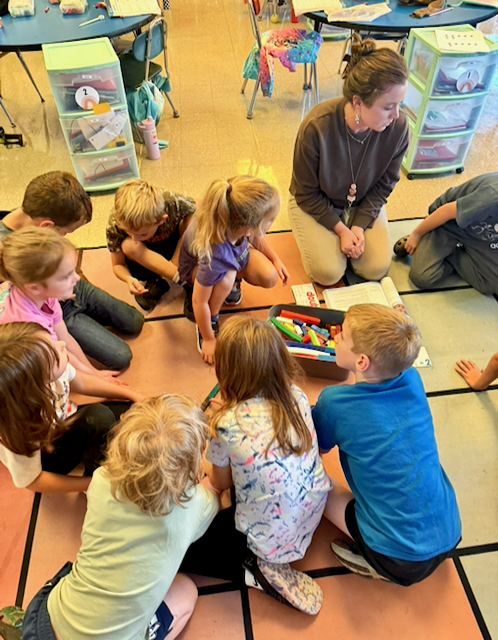 Elementary classroom scene showing approximately ten young students seated in a circle on a color-blocked classroom rug. An educator kneels among the group, leading a small-group lesson or discussion. In the center of the circle is an open bin filled with colorful markers and classroom supplies, along with instructional materials placed on the floor. Students are gathered closely around the materials and appear focused on the activity. The classroom contains student desks and chairs arranged around the perimeter, with rolling plastic storage drawers and personal classroom items visible nearby. The room is brightly lit with a warm, welcoming learning environment that emphasizes collaboration, engagement, and hands-on instruction.