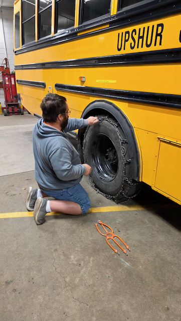 A transportation employee kneels beside a yellow school bus inside a vehicle maintenance garage while installing or adjusting tire chains on a rear wheel. The side of the bus displays the text "UPSHUR" in black lettering. Chain pliers are placed on the concrete floor near the wheel, and maintenance equipment is visible in the background of the garage. The employee is focused on securing the chains around the tire, demonstrating preventative maintenance and winter weather preparedness efforts that help ensure safe student transportation during adverse road conditions.