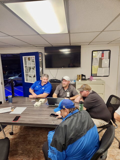 Five Upshur County Schools transportation employees sit around a conference table during a professional learning session. Several participants are looking at a laptop positioned in the center of the table as they review training materials. The meeting takes place in an office or transportation facility conference room with a wall-mounted television, filing cabinet, bulletin board, and office chairs visible in the background. One participant is seated in the foreground wearing a blue jacket and cap. The training focused on the AngelTrax school bus camera system, a safety technology used on district buses. The session was held during a countywide Professional Learning Day dedicated to staff development and school safety initiatives.