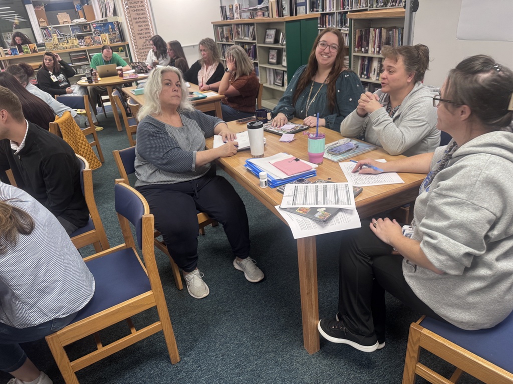 A group of educators and staff members participate in a professional learning session in a school library. Small groups are seated around wooden tables engaged in discussion and collaborative activities. Papers, notebooks, laptops, tablets, and drink containers are spread across the tables as participants review materials and share ideas. Bookshelves filled with books line the walls in the background, and additional staff members are working in groups throughout the room. The session focuses on student engagement and building systems of support that help students succeed academically and socially. The image captures active collaboration and professional development during a districtwide Professional Learning Day.