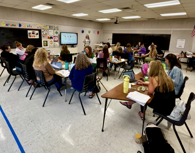 Educators participate in a professional learning session in a classroom setting. Several tables are arranged throughout the room with staff members seated in small groups reviewing materials, taking notes, and discussing instructional strategies. A presenter stands at the front of the room near a large display screen showing presentation content. Notebooks, instructional resources, laptops, and water bottles are visible on the tables. Classroom bulletin boards and educational displays line the walls. The professional development session focused on 95% Phonics and Tier II literacy intervention strategies to strengthen reading instruction and support student literacy growth.