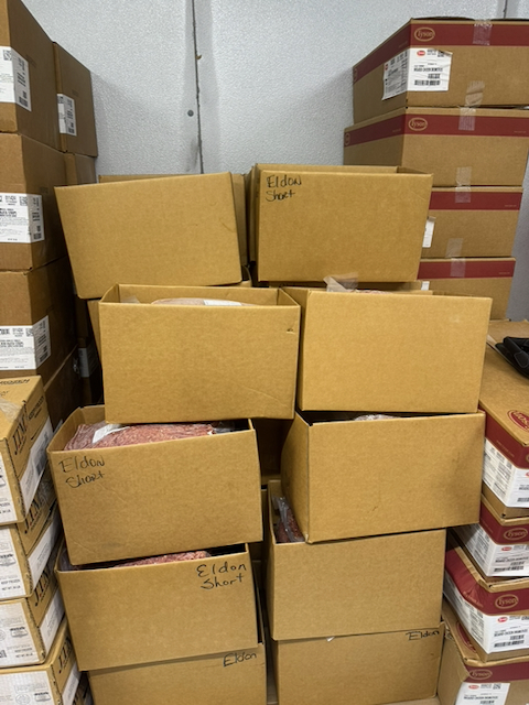 Stacks of open cardboard boxes filled with packaged ground beef are stored in a refrigerated food storage area. Several boxes are handwritten with the name "Eldon Short," identifying the local supplier. The boxes contain large sealed packages of ground beef stacked on pallets and shelves, with additional food service boxes visible along both sides of the storage room. The images document locally sourced ground beef purchased from Shorts Family Farm for use in the Upshur County Schools Child Nutrition Program.