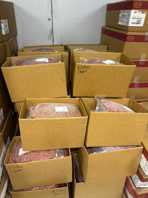 Stacks of open cardboard boxes filled with packaged ground beef are stored in a refrigerated food storage area. Several boxes are handwritten with the name "Eldon Short," identifying the local supplier. The boxes contain large sealed packages of ground beef stacked on pallets and shelves, with additional food service boxes visible along both sides of the storage room. The images document locally sourced ground beef purchased from Shorts Family Farm for use in the Upshur County Schools Child Nutrition Program.