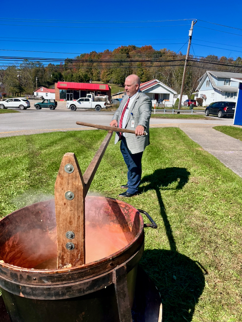 A large copper kettle of apple butter simmers outdoors on a grassy lawn, with steam rising from the surface. A person wearing a gray blazer, red tie, blue pants, and an identification badge stands beside the kettle, using a long wooden paddle attached to a wooden stirring mechanism. In the background are a roadway, several vehicles, utility poles and power lines, homes, and a tree-covered hillside displaying fall colors beneath a clear blue sky. The scene shows a traditional apple butter-making process taking place on a sunny autumn day.