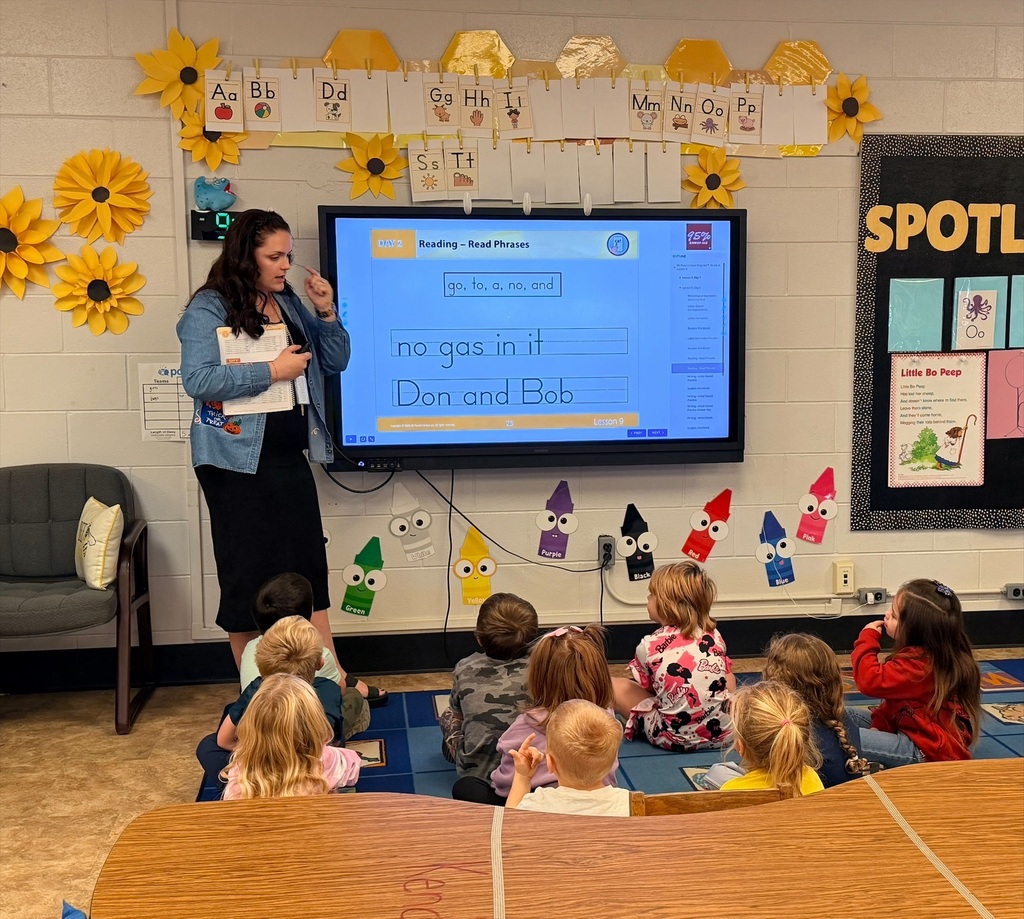 A teacher stands beside an interactive classroom display while leading a small-group reading lesson with elementary students seated on a blue carpet. The screen shows a phonics and reading exercise titled “Reading – Read Phrases,” with phrases including “no gas in it” and “Don and Bob.” Students face the display and teacher, participating in the lesson. The classroom features bright yellow sunflower decorations, an alphabet display hanging across the wall, colorful crayon-shaped cutouts labeled by color, and a literacy bulletin board displaying the nursery rhyme “Little Bo Peep.” The room is well-organized and designed to support early literacy instruction through direct teaching and student engagement.