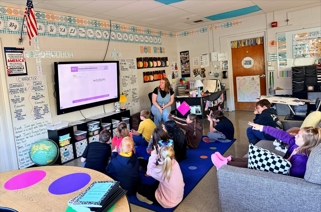 A classroom literacy lesson is underway as a teacher sits near an interactive display while approximately a dozen elementary students gather on a blue rug and classroom seating area. The display shows a reading lesson focused on multisyllabic words, with the word “wisdom” visible on the screen. Several students have their hands raised and appear to be participating in the discussion. The classroom walls are filled with educational materials, including an alphabet border, vocabulary and character trait charts, writing examples, classroom expectations, and student work displays. A sign reading “HOW WORK” hangs above a display of colorful student projects. An American flag hangs in the front corner of the room, and a globe sits beside a storage unit beneath the display screen. The classroom door is labeled “THIRD GRADE” and decorated with fall-themed artwork. Tables, books, flexible seating options, and organized learning materials surround the instructional space, creating an environment focused on active student engagement and literacy development