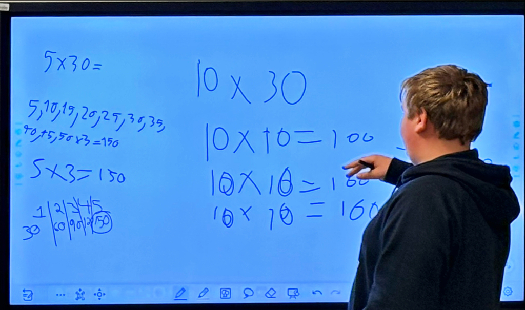 A fifth-grade student stands at an interactive classroom display demonstrating a multiplication problem to classmates. The screen contains handwritten math work showing multiple strategies for solving 5 × 30, including skip counting by fives (5, 10, 15, 20, 25, 30, 35), calculations involving 10 × 30, 10 × 10 = 100, and a number line illustrating multiples of 30 up to 150. The student points to the work on the display while explaining the solution process. The image highlights student-led learning, mathematical reasoning, and the use of visual problem-solving strategies to build understanding of multiplication concepts.