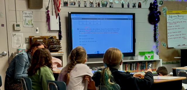 Students sit at a group table in an elementary classroom while participating in a math lesson displayed on a large interactive board at the front of the room. The screen shows a mathematics problem with answers and teacher annotations. Classroom bookshelves line the wall beneath the display, and educational posters, student work, and an American flag are visible around the room. Students face the board and engage with the lesson as mathematical concepts are reviewed and discussed.