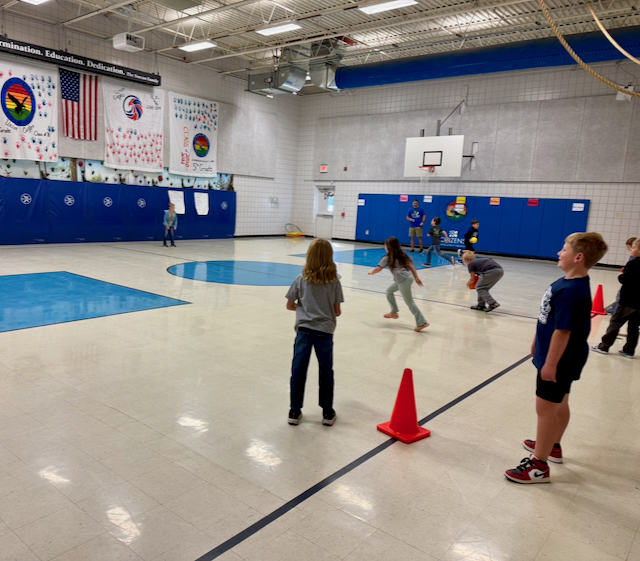 Students participate in a physical education activity inside a school gymnasium. Several students stand near orange cones while others run and throw balls across the gym floor. Blue wall padding, basketball hoops, and student-created banners decorate the space, with a teacher supervising the activity in the background.