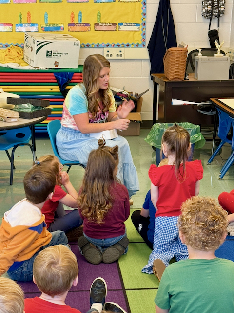 An elementary classroom gathers for a small-group lesson as students sit on a colorful carpet facing an instructor seated in a chair. The instructor leads a hands-on discussion using gestures while students listen and participate. Bright classroom materials, stacked gym mats, storage bins, and a bulletin board displaying the months of the year are visible in the background, creating an engaging early learning environment focused on interaction, communication, and foundational skill development.