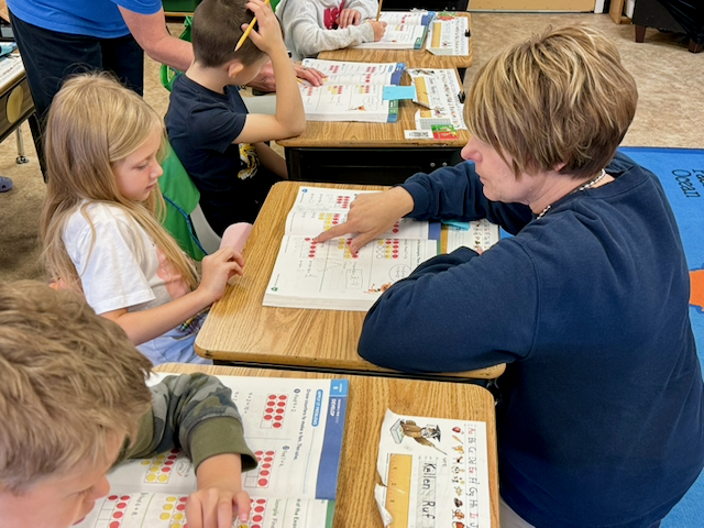 A building principal works one-on-one with an elementary student at a desk during a math lesson. The student and teacher review a workbook page featuring counting exercises and colored dot models while other students complete similar activities at nearby desks. Educational materials, number charts, and math workbooks are visible throughout the classroom, highlighting small-group instruction and individualized learning support.