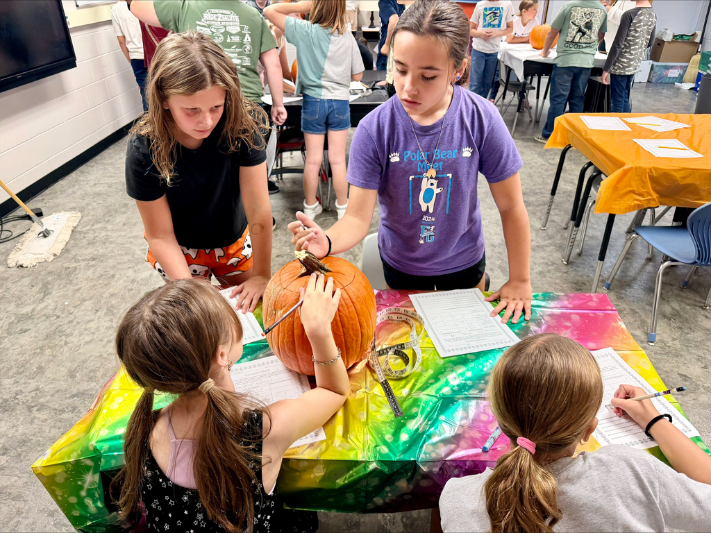 Four elementary students gather around a large pumpkin during a hands-on STEM activity in a classroom. The students use worksheets, measuring tools, and pencils to record observations and predictions about the pumpkin's weight, size, ridges, and seed count. Additional groups of students can be seen in the background working with pumpkins at other stations. The activity combines math, science, measurement, estimation, and data collection in a collaborative learning environment.