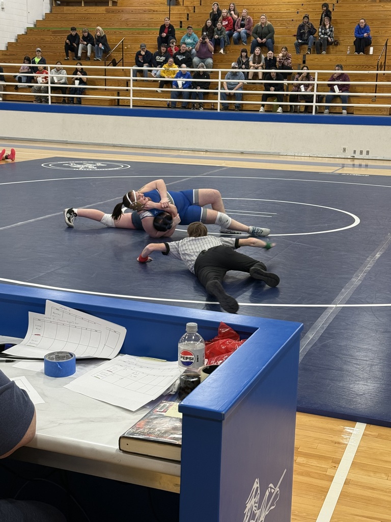 Three wrestlers are on the mat during a match. One lies on their back, another lies face down, and a referee is kneeling. Spectators sit on bleachers.