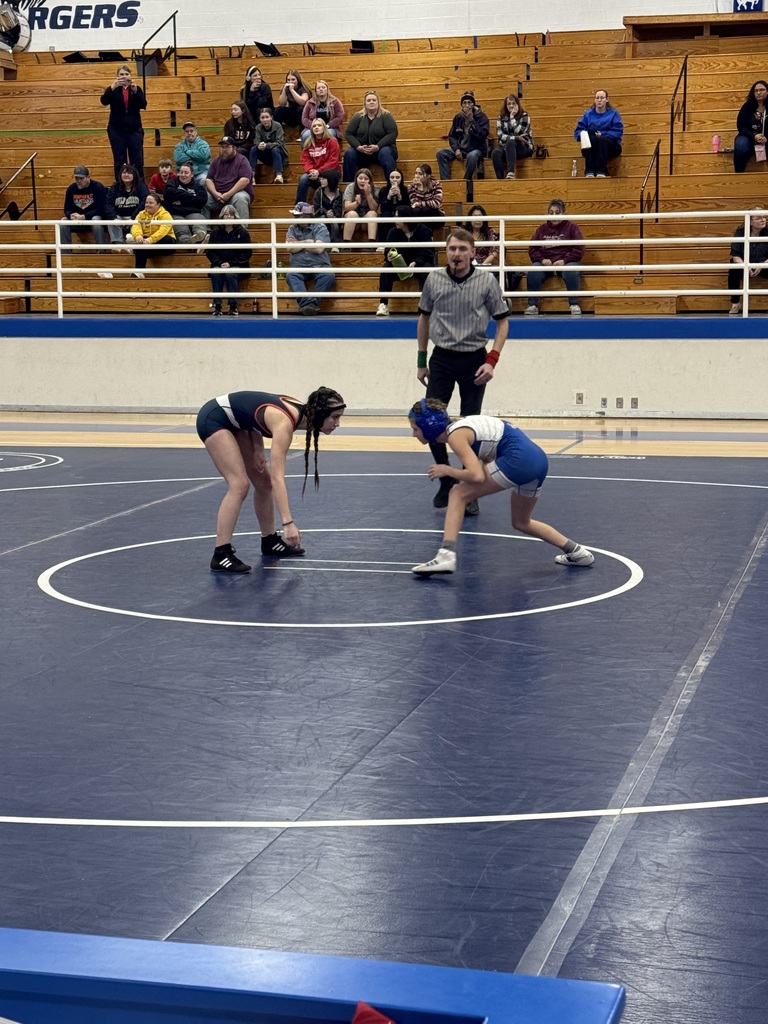 Two wrestlers in shorts, one with a blue helmet, compete in a ring while an official watches.