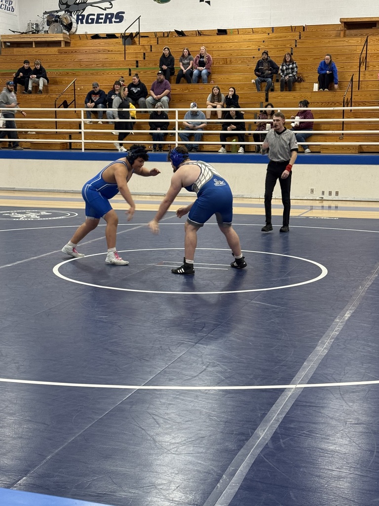 Two men in blue singlets compete in a wrestling match, surrounded by spectators in bleachers.