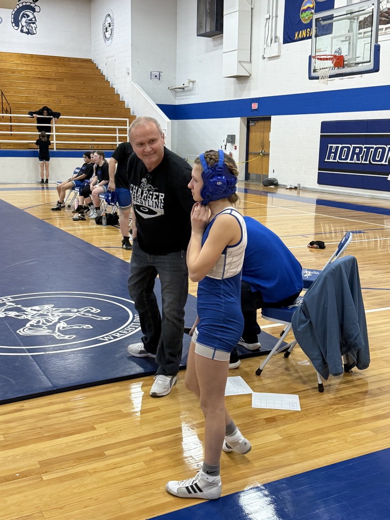 A man and a girl are standing on a blue mat in a gym. Spectators sit on bleachers.