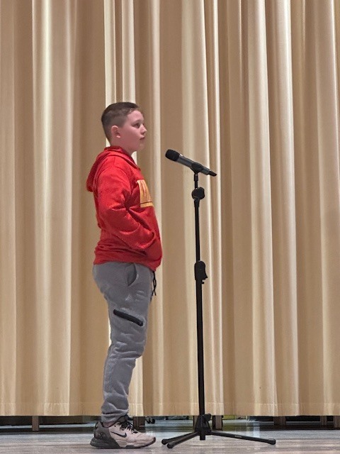 A young person stands on a stage, wearing a red hoodie and gray pants, speaking into a microphone.