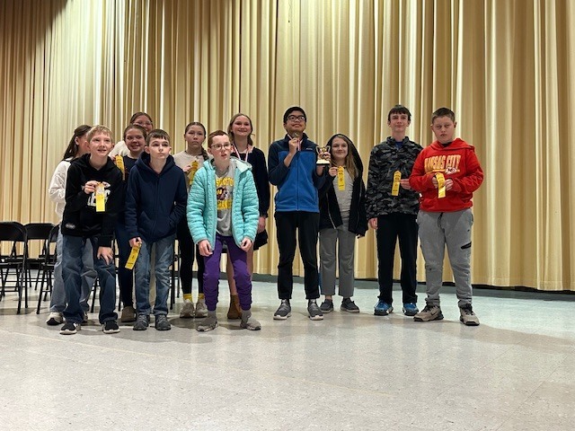 Eleven children, some holding certificates, stand in a row. The room has white walls and yellow curtains.