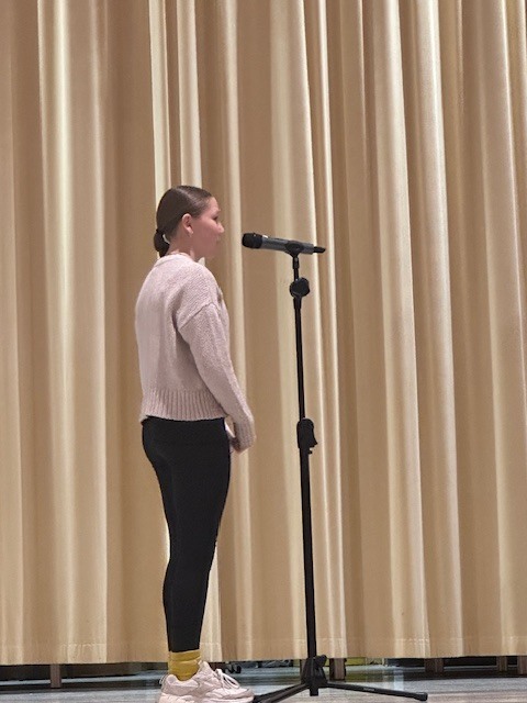 Person in white top and black pants stands at a microphone on a stage, with a curtain in the background.