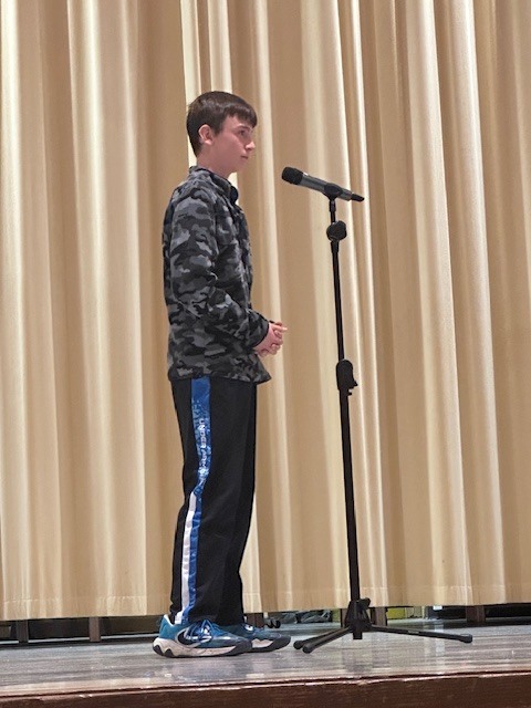 A person in a camouflage jacket and sneakers stands on a stage with a microphone.