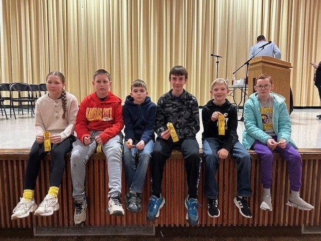 Group of children wearing winter clothing sitting on a stage, holding certificates. Behind them is a stage with a podium and curtains.